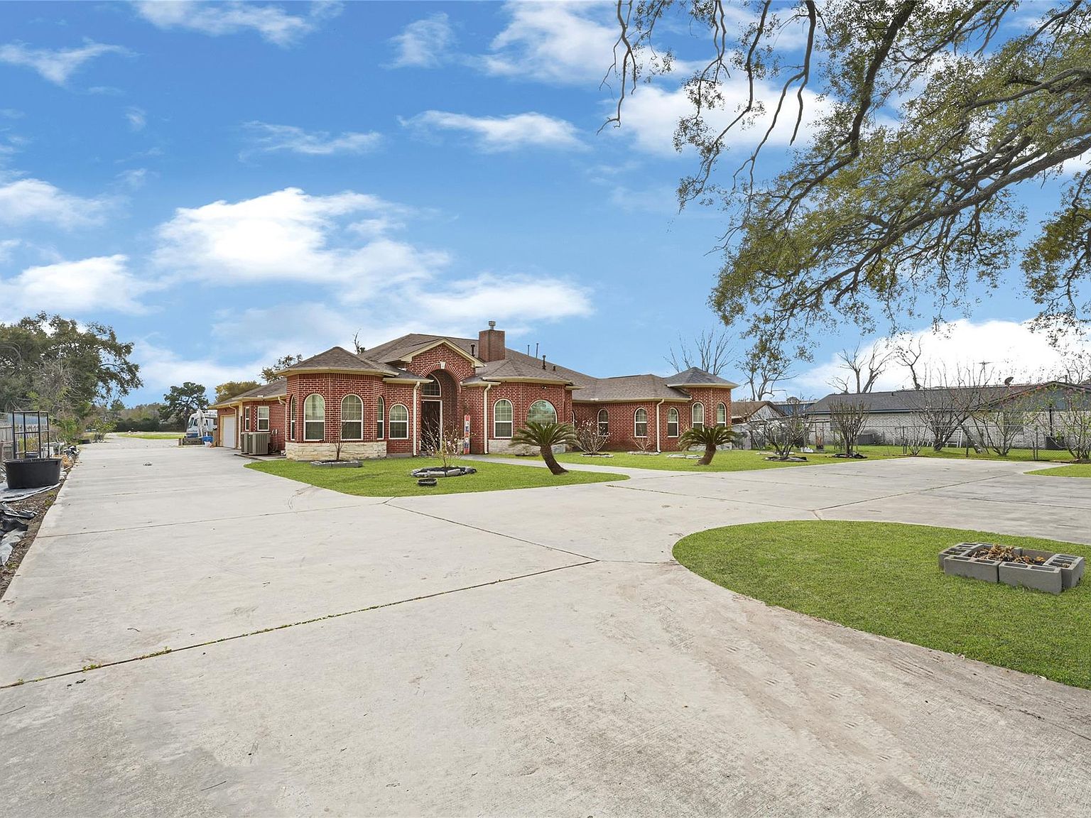 Wide three-quarter view of 6519 Breen showing the brick estate set back behind a circular concrete drive with palms and a mature oak in the foreground